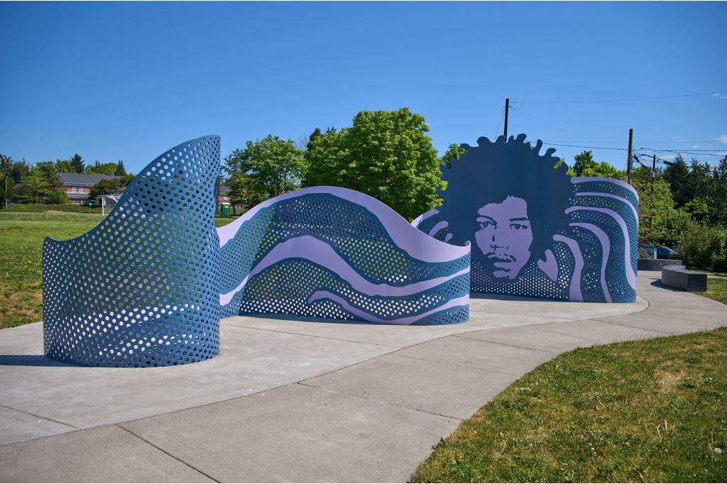 A wavy blue metal art installation in a park, featuring abstract perforations and a large cutout of a person's face with an afro, set against a backdrop of trees and a clear sky.