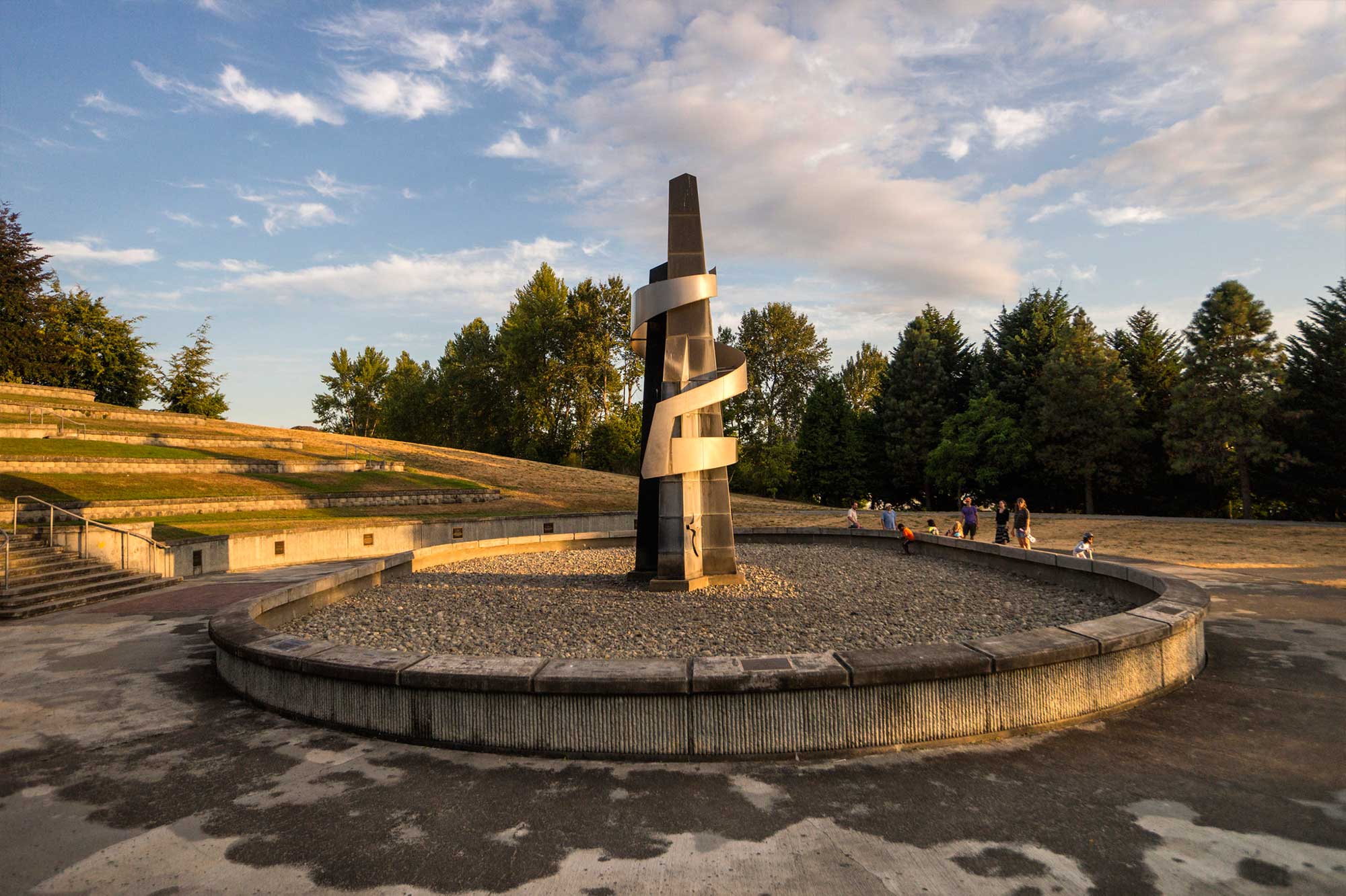 A tall metal sculpture with a twisting ribbon design stands in the center of a circular stone plaza surrounded by trees and grass, with people walking nearby under a partly cloudy sky.