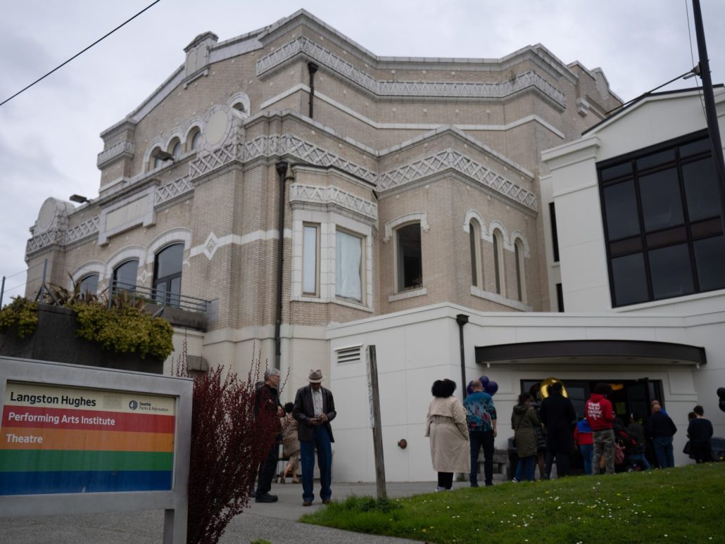 A group of people stands outside the historic Langston Hughes Performing Arts Institute, a large beige brick building. A sign in front identifies the location and theatre. The sky is overcast.