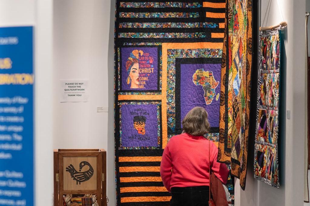 A person with gray hair and a red sweater views colorful, African-inspired quilts hanging on a wall in an exhibit, featuring portraits, patterns, and an outline of Africa. A "Do Not Touch" sign is visible nearby.
