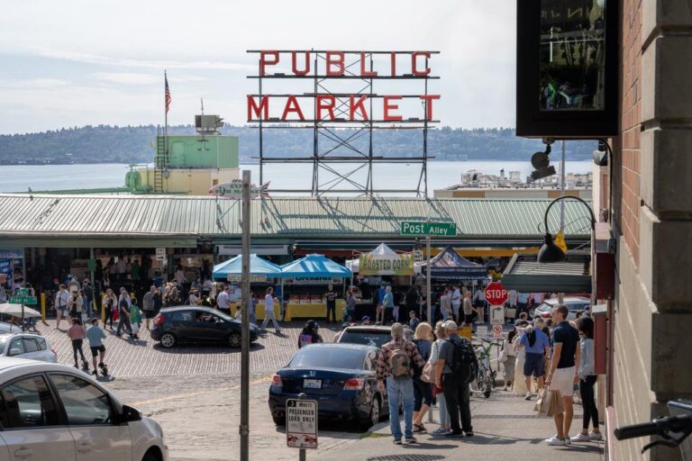 Crowds gather at Seattle's Pike Place Market beneath the iconic red "Public Market" sign, with vendor tents, cars, and a view of the water and hills in the background.