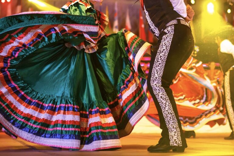 A woman in a vibrant, multicolored traditional Mexican dress dances beside a man in ornate black mariachi pants under bright, festive lights.