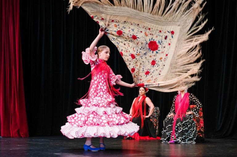 A young girl in a pink polka-dot flamenco dress and blue shoes dances on stage, waving a fringed shawl. Two women in black dresses with red scarves sit in the background, watching her performance.