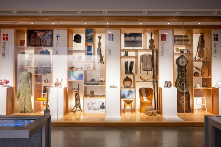 A museum display showcases traditional artifacts, clothing, and crafts from Denmark, Finland, Iceland, Norway, and Sweden, arranged in illuminated wooden shelves behind glass panels labeled with each country’s flag and name.