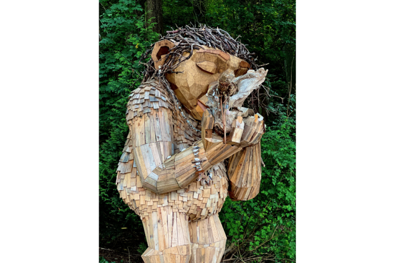 A large wooden sculpture of a troll-like creature stands in a forest, holding and examining a smaller wooden figure. The sculpture is made from various pieces of wood and branches, blending with the green foliage behind it.