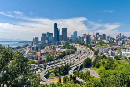 A view of a city skyline with many tall buildings, busy highways, green trees in the foreground, and water with distant mountains in the background under a blue, partly cloudy sky.