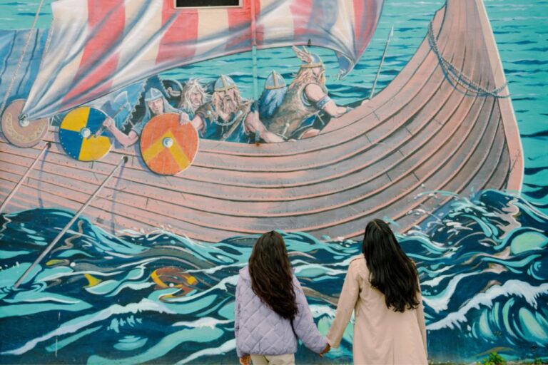 Two people with long dark hair, seen from behind, hold hands while looking at a colorful mural of Vikings rowing a ship on the ocean.