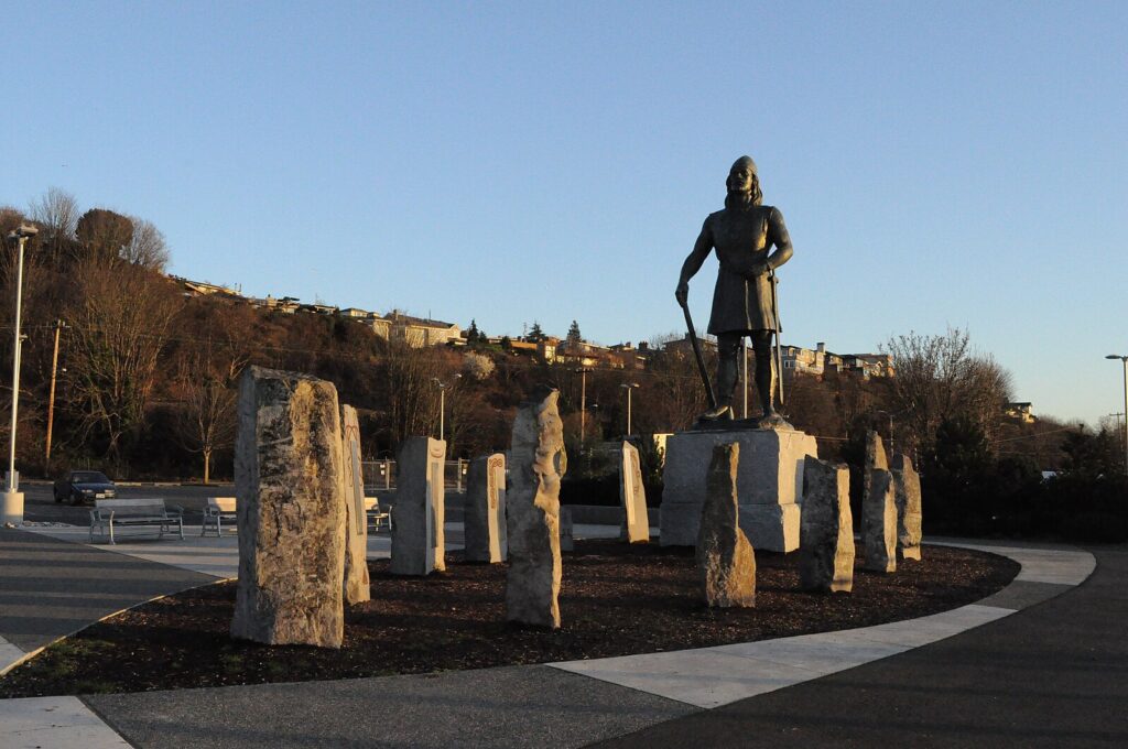 A statue of a Viking figure stands on a stone pedestal, surrounded by upright stones in a circular arrangement, with houses on a hillside in the background under clear blue sky.