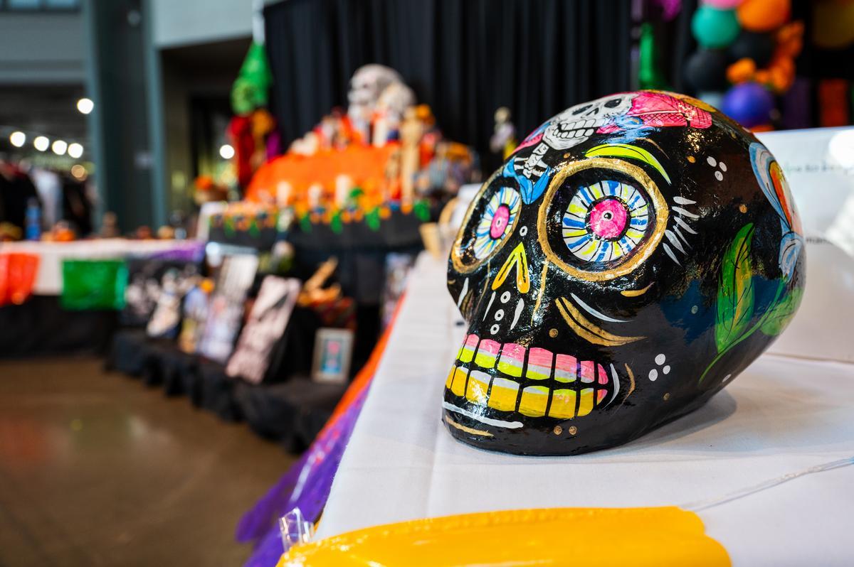 A brightly painted Day of the Dead skull sits on a table in the foreground, with a colorful Día de los Muertos altar, flowers, and photos of loved ones displayed in the background.