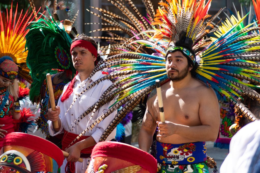 Two men wearing vibrant Aztec feather headdresses and colorful traditional attire stand in sunlight, holding percussion sticks, participating in a cultural dance or ceremony. Other dancers in similar outfits are visible in the background.