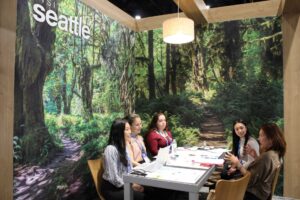 Five women sit around a table in a booth with forest-themed wall graphics and a “Seattle” sign, having a discussion with laptops, papers, and drinks in front of them.