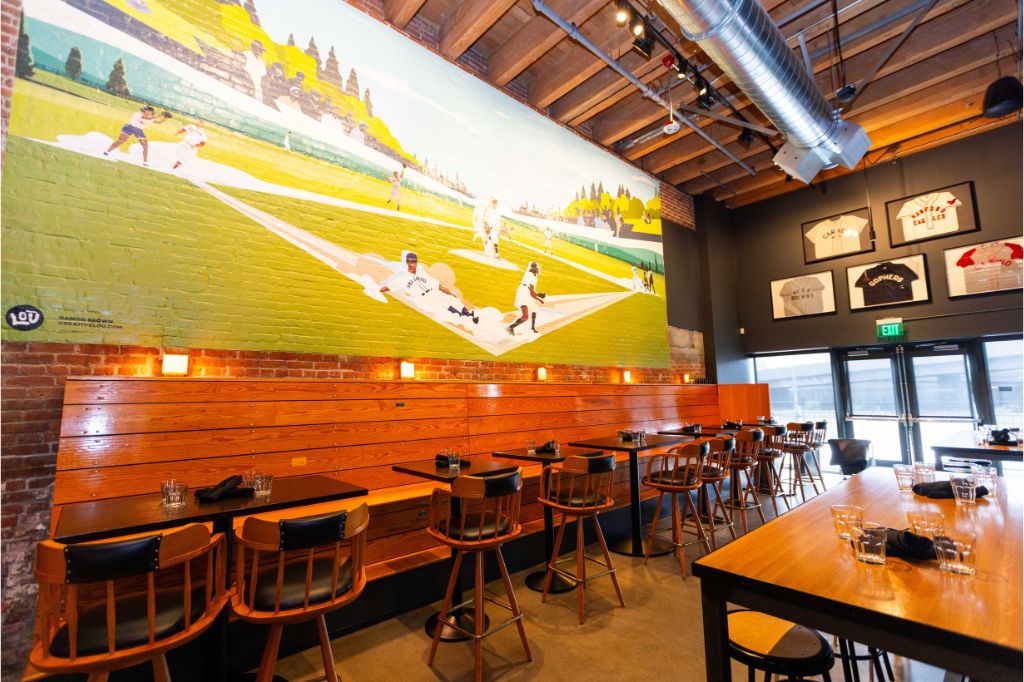 A restaurant with wooden tables and chairs, exposed ceiling beams, and a large mural of a baseball game on the wall. Lit candles line the wall, and framed sports jerseys hang above the entrance.
