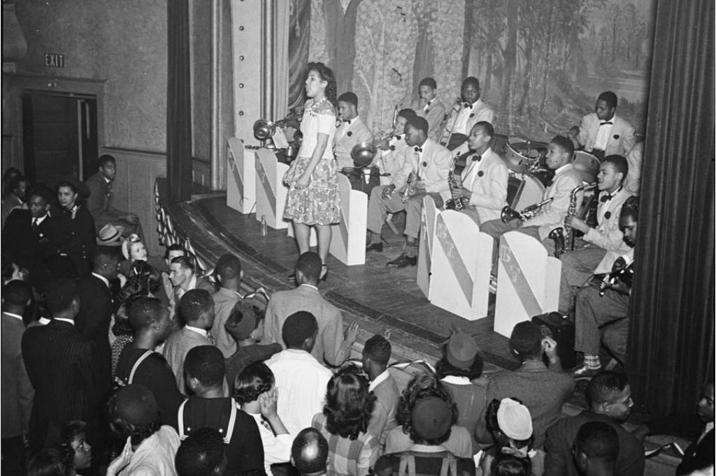 A female singer performs on stage with a big band while a large, well-dressed audience gathers in front, watching intently in a historic, possibly 1940s, jazz or swing club setting.