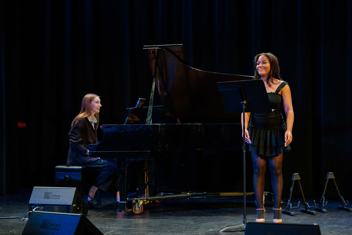 A woman sings at a microphone stand on stage while another woman plays a grand piano behind her. Both performers are dressed in black, and the background features dark curtains.