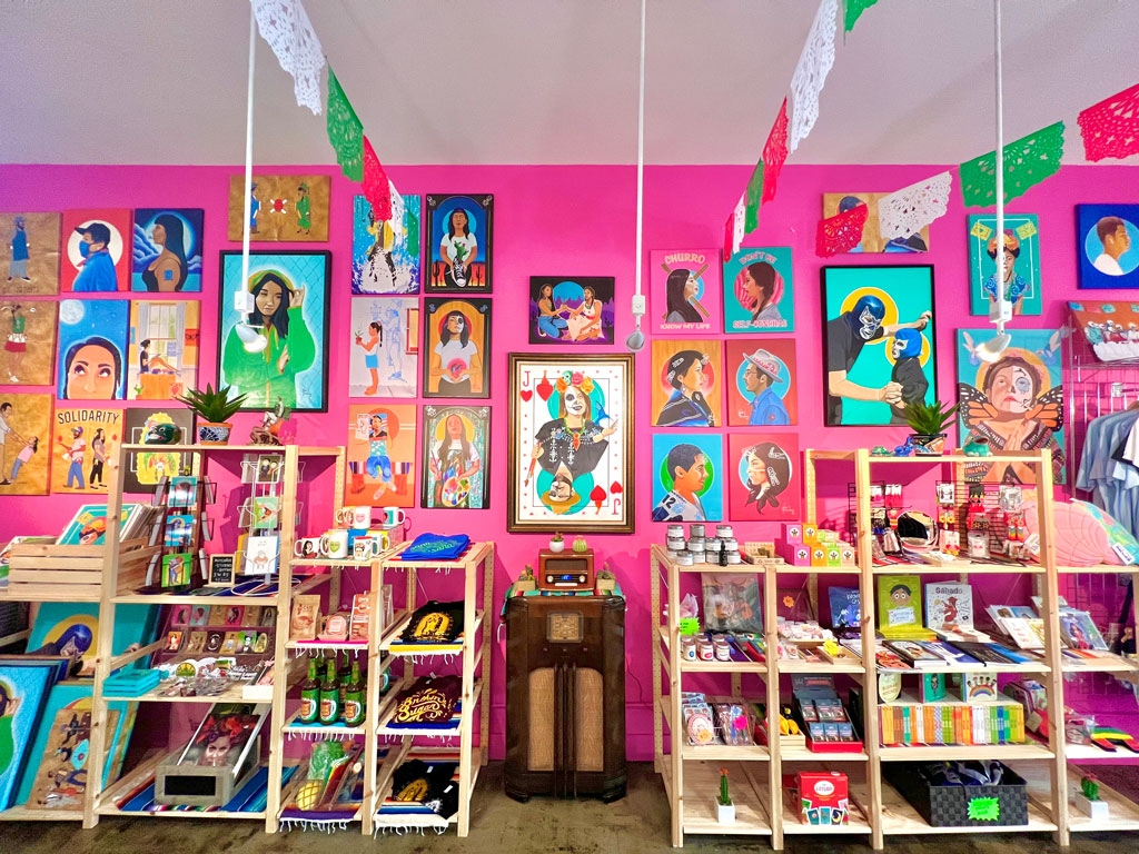 A colorful shop interior with shelves of books, gifts, and snacks. The bright pink wall is decorated with vibrant portraits of women. Traditional papel picado banners hang from the ceiling, adding to the festive atmosphere.