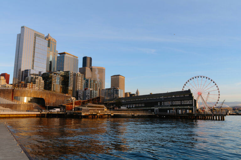 Seattle waterfront at sunset in 2026, featuring the city skyline, a large Ferris wheel, and a pier with buildings beautifully reflected in the calm water.