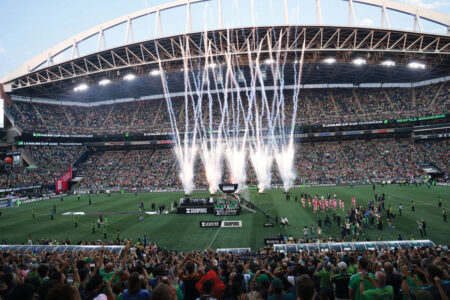 A large stadium filled with cheering fans as fireworks shoot into the sky from a stage on the soccer field during a post-match celebration. Photographers and players gather near the stageand the stands are packed with people.