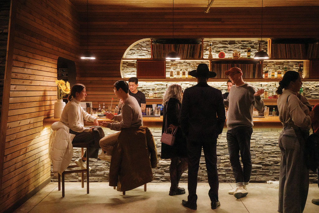 A cozy, warmly lit bar with wooden walls and shelves. People are chatting and enjoying drinks; some stand at the bar while a couple sits at a high table. Two bartenders work behind the counter.