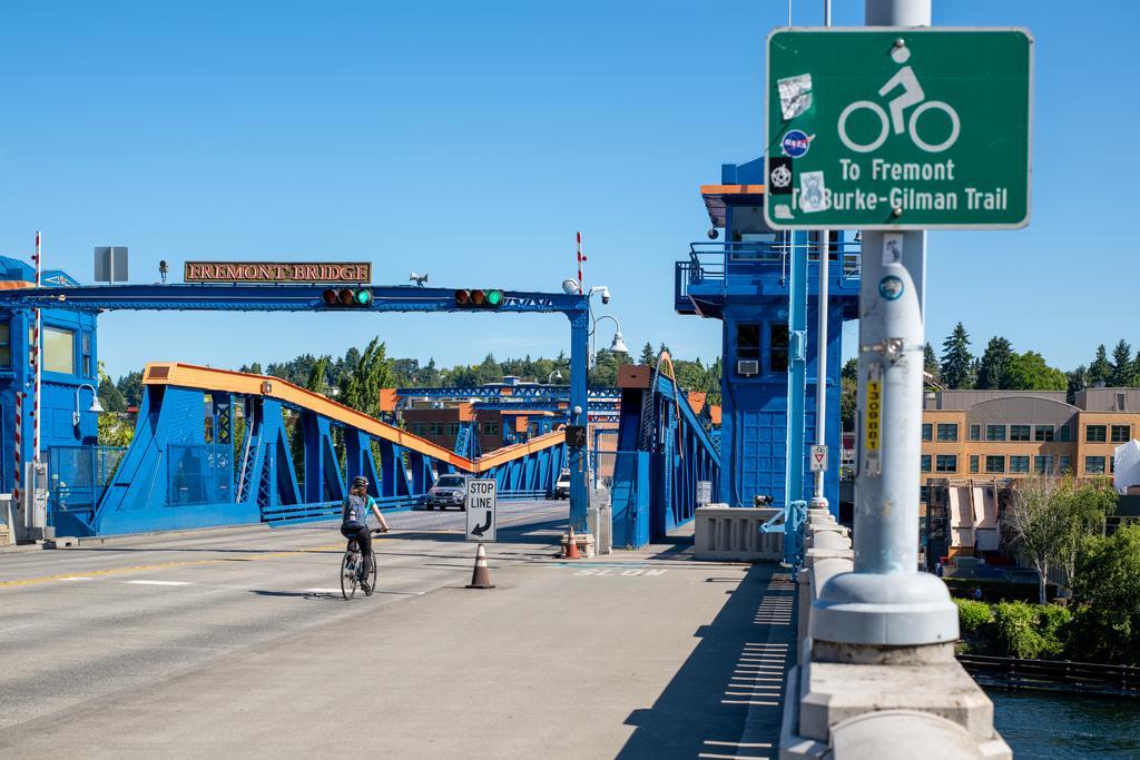 A cyclist rides across the blue Fremont Bridge in Seattle on a sunny day. A green sign indicates directions to Fremont and the Burke-Gilman Trail for bicyclists. Trees and buildings are visible in the background.