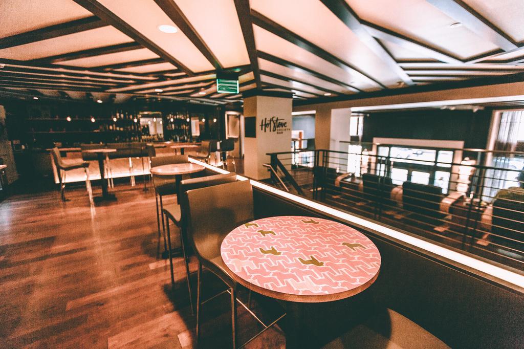 A modern, stylish restaurant interior with wooden floors, patterned tables, cushioned chairs, and a bar area in the background. Soft lighting and a railing overlook the space below.