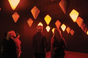 Four people stand in a dark room illuminated by many orange, diamond-shaped hanging lanterns, gazing upward at the glowing lights—a striking example of immersive Seattle Art with a warm and contemplative atmosphere.