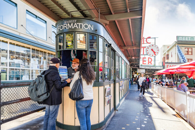 People stand at an information booth at Pike Place Market in Seattle. The lively scene, featured in the Seattle Monthly Resource Guide, includes shoppers, outdoor seating, and the iconic “Public Market Center” sign with its large red clock.
