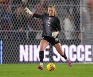 A soccer goalkeeper in a black uniform with the number 18 kicks a multicolored ball during a Seattle sports game. The goal net and 