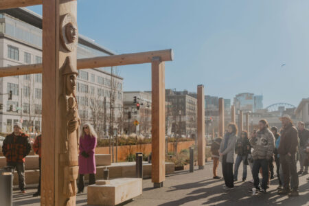 A group of people gather outdoors near wooden totem poles in a city plaza on a sunny day, with modern buildings and a blue sky in the background.