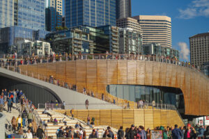 Crowds of people walk and gather on and around a modern, curved wooden structure with city skyscrapers in the background under a blue sky.
