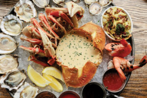 A seafood platter with oysters, crab legs, shrimp, lemon wedges, dipping sauces, and a crab shell filled with creamy sauce, all served on a bed of ice.