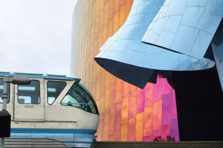 A white and blue monorail passes in front of a modern building with curved, metallic panels in blue, gold, and pink tones.