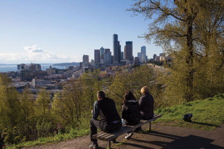 Three people sit on a bench overlooking Seattle’s skyline with tall buildings under a clear sky, surrounded by trees and greenery, on a sunny day.
