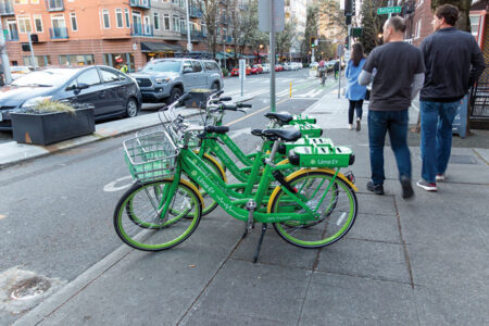 Three green Lime-E electric bikes are parked on a city sidewalk near a bike lane. People walk by, and cars are parked along the street with apartment buildings in the background.