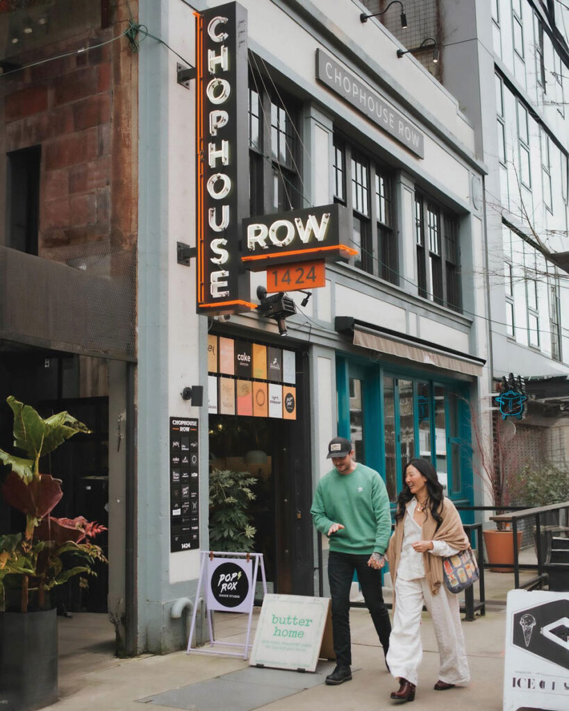 A stylish couple walks past the entrance of Chophouse Row, an urban building with neon signage and various shop signs, potted plants, and outdoor seating visible.