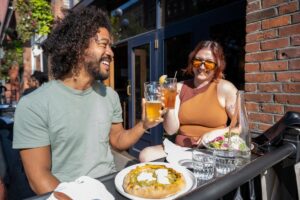 Two people sitting outdoors at a restaurant table, smiling and clinking glasses of beer. There’s a pizza, salad, and glasses of water on the table, with a brick building in the background on a sunny day.