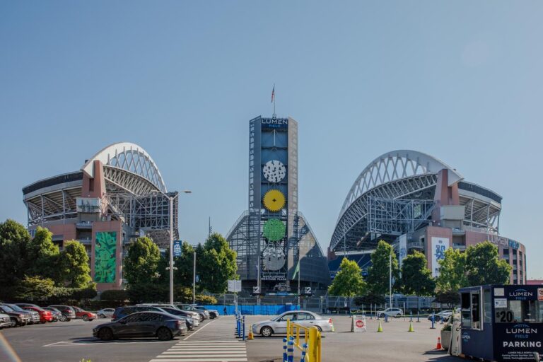 Seattle Stadium features a large arched roof and a central clock tower, standing behind a parking lot filled with cars on a sunny day. Trees and signs surround the entrance, enhancing the welcoming atmosphere.