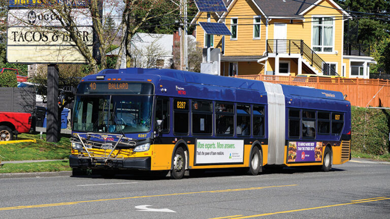 A blue and yellow articulated city bus marked "40 Ballard" drives down a street with houses and a taco restaurant in the background on a sunny day.