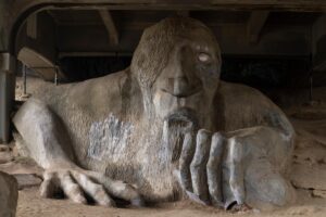 A large concrete sculpture of a troll with long hair and one visible eye sits under a bridge, clutching a real Volkswagen Beetle in its left hand. The sculpture appears weathered and textured.