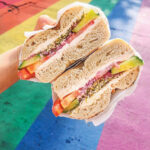A hand holds a sliced bagel sandwich filled with cream cheese, tomato, cucumber, and seasoning. The background features a vibrant rainbow-striped crosswalk.