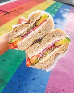 A hand holds a sliced bagel sandwich filled with cream cheese, tomato, cucumber, and seasoning. The background features a vibrant rainbow-striped crosswalk.
