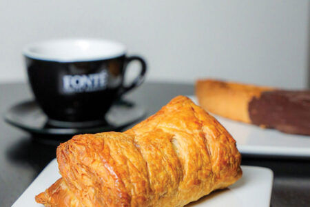 A flaky, golden pastry sits on a white square plate, with a black coffee cup and saucer and a chocolate-dipped pastry in the background on a dark table.