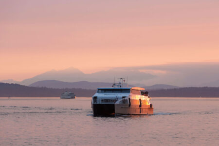 A ferry and a tour boat travel on calm water at sunset, with orange-pink skies and silhouettes of distant mountains and trees in the background.
