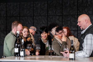 A group of six adults sit at a table with wine bottles and glasses, laughing and enjoying a lively conversation in a modern, dark-toned space.