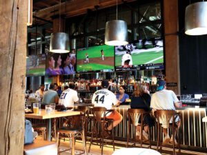 People sit at a bar watching sports on large TVs. The bar has wooden beams, shelves of bottles, and hanging lights. Some patrons wear sports jerseys, and drinks are on the counter. The atmosphere is casual and lively.