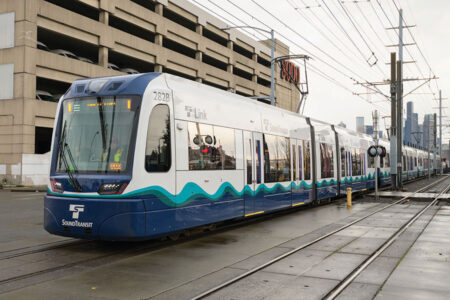 A blue and white Sound Transit Link light rail train is stopped on wet tracks near a multi-story parking garage in an urban area, with overhead power lines and city buildings visible in the background.