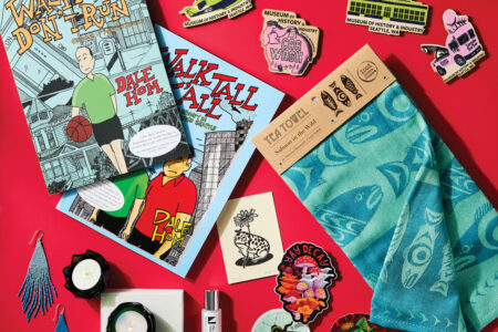 A flat lay on a red background displays two comic books, colorful embroidered patches, a blue patterned tea towel, blue feather earrings, a small candle, a spray vial, and a few vibrant stickers.