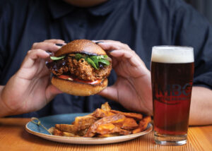 A person holds a fried chicken sandwich with lettuce and tomato, next to a plate of fries and a glass of dark beer on a wooden table.