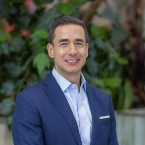 A man in a blue suit jacket and light blue dress shirt stands smiling in front of a background of lush green plants and pink flowers.