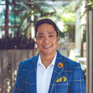 A man in a blue checked blazer with a yellow pocket square and flower pin smiles while standing outdoors in a lush, green, modern garden setting.