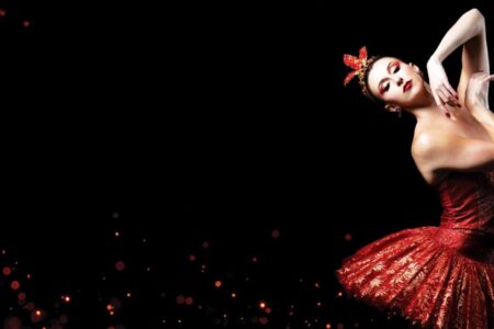 A ballerina in a red and gold tutu poses gracefully against a black background, with red and gold glowing particles scattered around her.